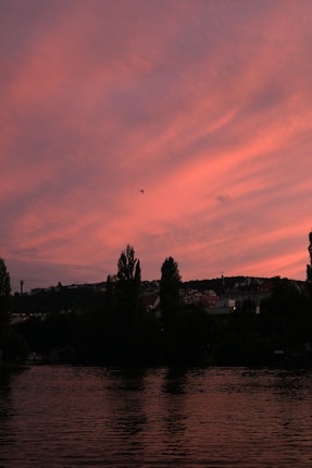 A serene landscape at dusk featuring a river with slight ripples, silhouetted trees lining the riverbank, and a vast sky filled with soft pink and orange clouds. A small bird is visible flying across the sky.