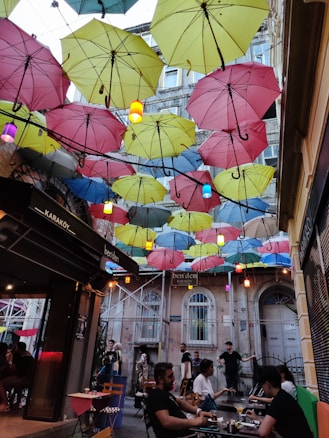 A vibrant street scene with colorful umbrellas hanging overhead, creating a visually striking canopy. People are seated at outdoor tables enjoying their time, set against a backdrop of aged buildings. Warm light from small hanging lamps complements the lively atmosphere.
