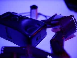 A vintage film camera rests on a surface, surrounded by several strips of undeveloped film negatives. The lighting is a moody purple and blue, creating an atmospheric scene.