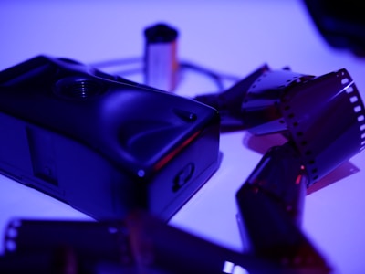 A vintage film camera rests on a surface, surrounded by several strips of undeveloped film negatives. The lighting is a moody purple and blue, creating an atmospheric scene.