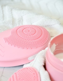A close-up of a pink silicone facial cleansing brush with fine bristles arranged in a circular pattern. The brush is resting on a soft, white textured towel. Nearby, there's an open jar of skincare product with a pink lid.