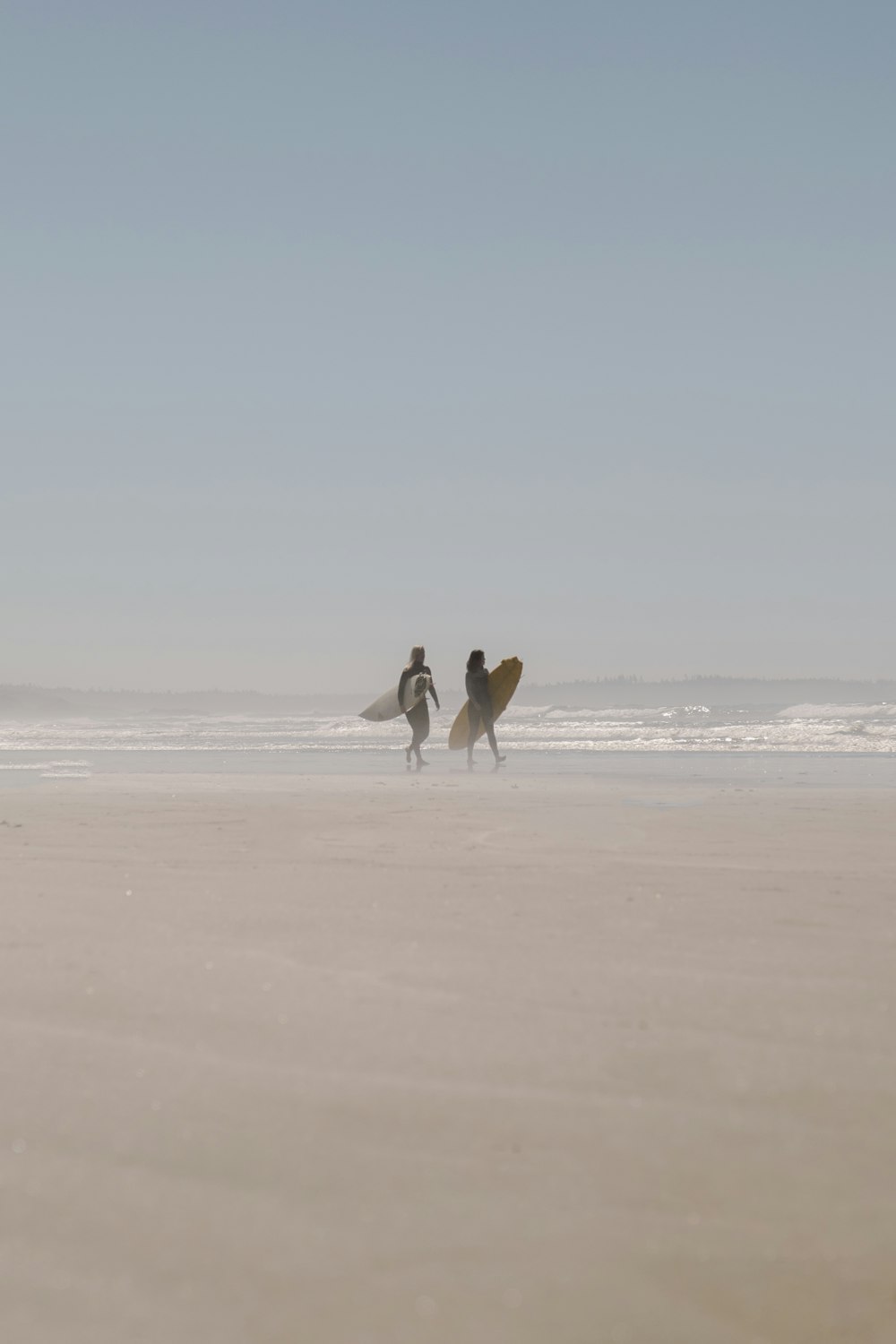 흰색 셔츠와 검은 바지를 입은 남자가 낮에 해변을 걷는 흰색 서핑보드를 들고 있다