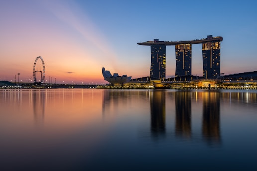 A stunning skyline at dusk featuring the Marina Bay Sands hotel complex with its iconic three towers topped by a sky park. The calm water in the foreground reflects the buildings and the lights magnificently. To the left, the Singapore Flyer ferris wheel is visible against a colorful, serene sunset backdrop.