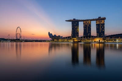 A stunning skyline at dusk featuring the Marina Bay Sands hotel complex with its iconic three towers topped by a sky park. The calm water in the foreground reflects the buildings and the lights magnificently. To the left, the Singapore Flyer ferris wheel is visible against a colorful, serene sunset backdrop.