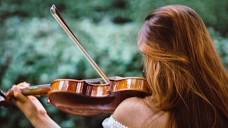 A person with long hair is playing a violin outdoors, with a blurred green background, suggesting a natural setting.