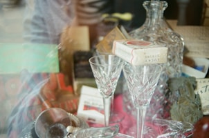 A collection of vintage items including a large crystal decanter, two crystal glasses, a pack of Lucky Strike cigarettes, and various books or boxes. The setting suggests an eclectic mix, possibly on display in a shop window.