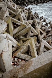 Triangular concrete structures are piled up along a rocky shoreline. The rough texture of these geometric shapes contrasts with the foamy waves of the sea. The scene has an industrial and rugged feel due to the arrangement of massive concrete blocks.