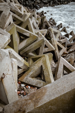 Triangular concrete structures are piled up along a rocky shoreline. The rough texture of these geometric shapes contrasts with the foamy waves of the sea. The scene has an industrial and rugged feel due to the arrangement of massive concrete blocks.