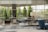 A spacious, modern lobby area with floor-to-ceiling windows allowing natural light to fill the space. The room is furnished with a mix of blue and gray upholstered chairs and sofas, along with a wooden tripod floor lamp. There are potted plants placed strategically, adding a touch of greenery. Outside the windows, manicured trees and a glimpse of parked cars can be seen.