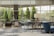 A spacious, modern lobby area with floor-to-ceiling windows allowing natural light to fill the space. The room is furnished with a mix of blue and gray upholstered chairs and sofas, along with a wooden tripod floor lamp. There are potted plants placed strategically, adding a touch of greenery. Outside the windows, manicured trees and a glimpse of parked cars can be seen.