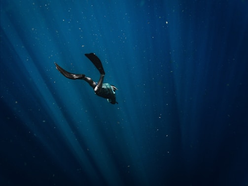 A solitary diver is gracefully swimming underwater, surrounded by soft, deep blue rays of light piercing through the water. The scene creates a sense of peace and wonder in the vast, open ocean.