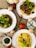 A variety of dishes are artistically arranged on a textured burlap surface. The focus is on three plates featuring different meals: one with stir-fried broccoli, another with pasta topped with a rich mushroom and tomato sauce, and the last with plain yellow spaghetti garnished with fresh basil leaves. Surrounding the plates are fresh ingredients including whole tomatoes, green chili peppers, and mushrooms. A small bowl of pesto sauce and a dish of grated cheese add to the culinary presentation.