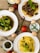 A variety of dishes are artistically arranged on a textured burlap surface. The focus is on three plates featuring different meals: one with stir-fried broccoli, another with pasta topped with a rich mushroom and tomato sauce, and the last with plain yellow spaghetti garnished with fresh basil leaves. Surrounding the plates are fresh ingredients including whole tomatoes, green chili peppers, and mushrooms. A small bowl of pesto sauce and a dish of grated cheese add to the culinary presentation.