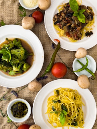 A variety of dishes are artistically arranged on a textured burlap surface. The focus is on three plates featuring different meals: one with stir-fried broccoli, another with pasta topped with a rich mushroom and tomato sauce, and the last with plain yellow spaghetti garnished with fresh basil leaves. Surrounding the plates are fresh ingredients including whole tomatoes, green chili peppers, and mushrooms. A small bowl of pesto sauce and a dish of grated cheese add to the culinary presentation.