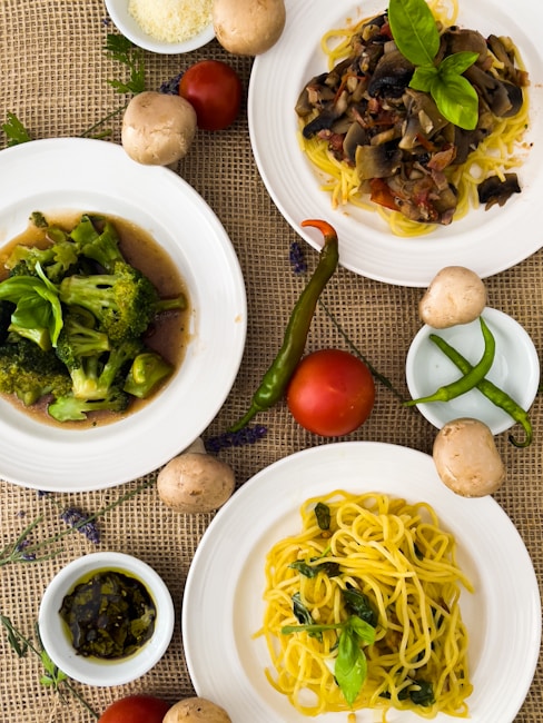A variety of dishes are artistically arranged on a textured burlap surface. The focus is on three plates featuring different meals: one with stir-fried broccoli, another with pasta topped with a rich mushroom and tomato sauce, and the last with plain yellow spaghetti garnished with fresh basil leaves. Surrounding the plates are fresh ingredients including whole tomatoes, green chili peppers, and mushrooms. A small bowl of pesto sauce and a dish of grated cheese add to the culinary presentation.