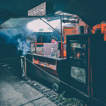 A vintage Hindustan Ambassador transformed into a fiery street BBQ cart with smoke rising against a gritty urban backdrop.