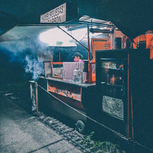 A street food cart illuminated under dim lighting, with smoke billowing from the cooking area. A vendor wearing a cap and mask is attending to the cart. Signs in Spanish advertise the food being served. The ambient atmosphere suggests an urban, possibly evening setting.