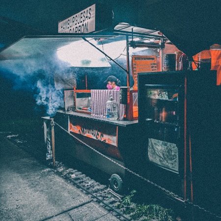 A street food cart illuminated under dim lighting, with smoke billowing from the cooking area. A vendor wearing a cap and mask is attending to the cart. Signs in Spanish advertise the food being served. The ambient atmosphere suggests an urban, possibly evening setting.