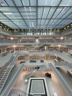 Five levels of open shelving filled with books line the walls of a large, modern library. The central area is open, with multiple floors visible, and a network of stairs and walkways connecting them. Sitting areas with small groups of people are scattered throughout, and bright natural light comes in through a large grid-patterned skylight above.