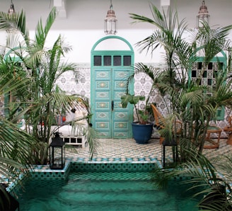 A serene courtyard featuring lush green plants surrounding a small pool of turquoise water, with a large ornate turquoise door set against a patterned ceramic tile wall. The furnishings include wooden chairs and a couch, with hanging lanterns adding to the ambiance.