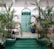 A serene courtyard featuring lush green plants surrounding a small pool of turquoise water, with a large ornate turquoise door set against a patterned ceramic tile wall. The furnishings include wooden chairs and a couch, with hanging lanterns adding to the ambiance.