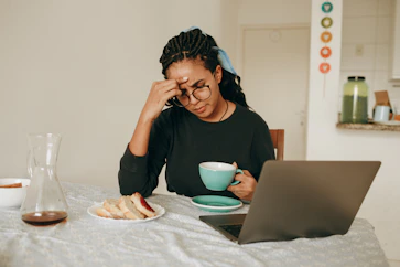 A tired woman sitting at a table with a laptop and a cup, resting her hand on her forehead.