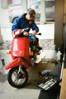 A person in a blue jacket and brown cap is working on a scooter inside a garage or workshop. The scooter is red and has missing parts, indicating it's being repaired or assembled. Nearby, there are tools scattered on the floor including a toolbox and a spray can.