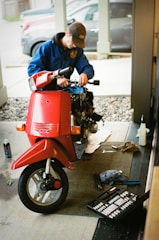 A person in a blue jacket and brown cap is working on a scooter inside a garage or workshop. The scooter is red and has missing parts, indicating it's being repaired or assembled. Nearby, there are tools scattered on the floor including a toolbox and a spray can.