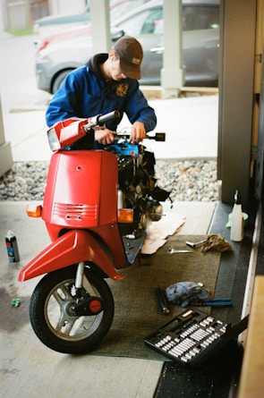 A person in a blue jacket and brown cap is working on a scooter inside a garage or workshop. The scooter is red and has missing parts, indicating it's being repaired or assembled. Nearby, there are tools scattered on the floor including a toolbox and a spray can.