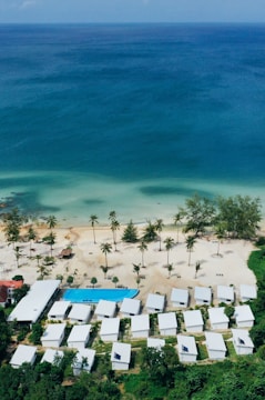 Aerial view of a seaside resort featuring a row of white-roofed buildings nestled among lush greenery. The clear, turquoise ocean borders a sandy beach dotted with palm trees. A rectangular swimming pool is situated near the beach, adding to the tranquil coastal setting.