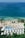 Aerial view of a seaside resort featuring a row of white-roofed buildings nestled among lush greenery. The clear, turquoise ocean borders a sandy beach dotted with palm trees. A rectangular swimming pool is situated near the beach, adding to the tranquil coastal setting.