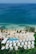 Aerial view of a seaside resort featuring a row of white-roofed buildings nestled among lush greenery. The clear, turquoise ocean borders a sandy beach dotted with palm trees. A rectangular swimming pool is situated near the beach, adding to the tranquil coastal setting.