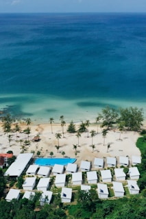 Aerial view of a seaside resort featuring a row of white-roofed buildings nestled among lush greenery. The clear, turquoise ocean borders a sandy beach dotted with palm trees. A rectangular swimming pool is situated near the beach, adding to the tranquil coastal setting.