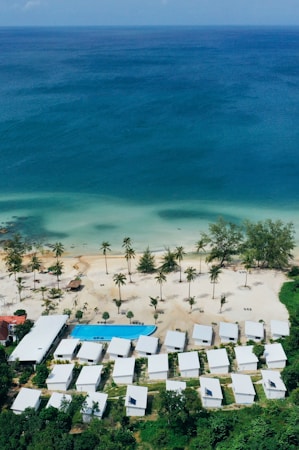 Aerial view of a seaside resort featuring a row of white-roofed buildings nestled among lush greenery. The clear, turquoise ocean borders a sandy beach dotted with palm trees. A rectangular swimming pool is situated near the beach, adding to the tranquil coastal setting.