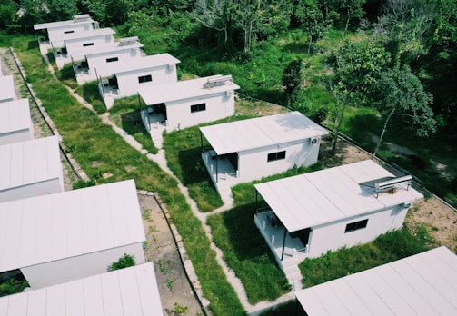 Aerial view of a series of small, white, rectangular houses with flat roofs arranged in rows. These homes are surrounded by lush greenery and grass-covered paths. Solar panels are visible on some of the rooftops.