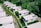Aerial view of a series of small, white, rectangular houses with flat roofs arranged in rows. These homes are surrounded by lush greenery and grass-covered paths. Solar panels are visible on some of the rooftops.