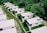 Aerial view of a series of small, white, rectangular houses with flat roofs arranged in rows. These homes are surrounded by lush greenery and grass-covered paths. Solar panels are visible on some of the rooftops.