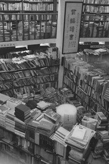 Black and white image of a cluttered bookstore filled with stacks of books on shelves and tables. Narrow aisles are formed between the piles of books, and a piece of scroll art with Asian calligraphy is prominently displayed on a central pillar.