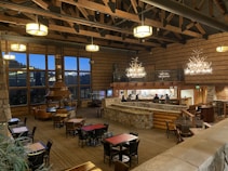 A spacious, cozy lodge with wooden beams and stone walls. It features large windows showcasing an evening view. The room includes a central fireplace with a copper hood and several seating areas, with tables and chairs arranged throughout. There is a bar on one side with patrons seated and a stone and wood design complementing the overall rustic aesthetic. Antler chandeliers hang from the ceiling, providing warm lighting.