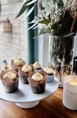 Cupcakes with creamy frosting and sprinkled toppings are neatly arranged on a white cake stand. A tall glass vase holds a bouquet of green leaves and white flowers next to a lit candle in a white jar.