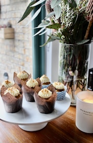 Cupcakes with creamy frosting and sprinkled toppings are neatly arranged on a white cake stand. A tall glass vase holds a bouquet of green leaves and white flowers next to a lit candle in a white jar.