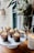 Cupcakes with creamy frosting and sprinkled toppings are neatly arranged on a white cake stand. A tall glass vase holds a bouquet of green leaves and white flowers next to a lit candle in a white jar.