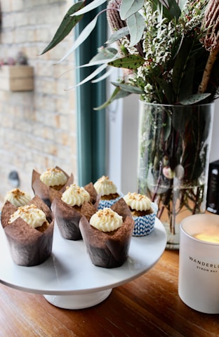 Cupcakes with creamy frosting and sprinkled toppings are neatly arranged on a white cake stand. A tall glass vase holds a bouquet of green leaves and white flowers next to a lit candle in a white jar.
