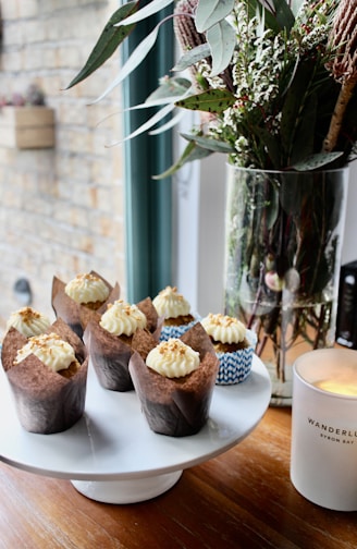 Cupcakes with creamy frosting and sprinkled toppings are neatly arranged on a white cake stand. A tall glass vase holds a bouquet of green leaves and white flowers next to a lit candle in a white jar.