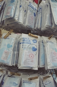 Multiple packages of face masks are hanging, each containing multiple masks labeled with information about their features. The packages are clipped with wooden clothespins and display text in different languages.