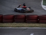 A go-kart zooms around a racetrack, with a driver wearing a helmet and racing suit. Red and black tires are stacked as barriers along the track. The asphalt is dark, and the grass is visible along the edge.