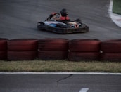 A go-kart zooms around a racetrack, with a driver wearing a helmet and racing suit. Red and black tires are stacked as barriers along the track. The asphalt is dark, and the grass is visible along the edge.