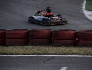 A go-kart zooms around a racetrack, with a driver wearing a helmet and racing suit. Red and black tires are stacked as barriers along the track. The asphalt is dark, and the grass is visible along the edge.