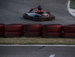 A go-kart zooms around a racetrack, with a driver wearing a helmet and racing suit. Red and black tires are stacked as barriers along the track. The asphalt is dark, and the grass is visible along the edge.