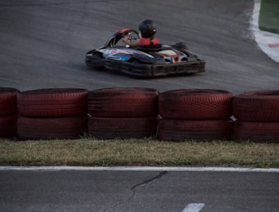 A go-kart zooms around a racetrack, with a driver wearing a helmet and racing suit. Red and black tires are stacked as barriers along the track. The asphalt is dark, and the grass is visible along the edge.